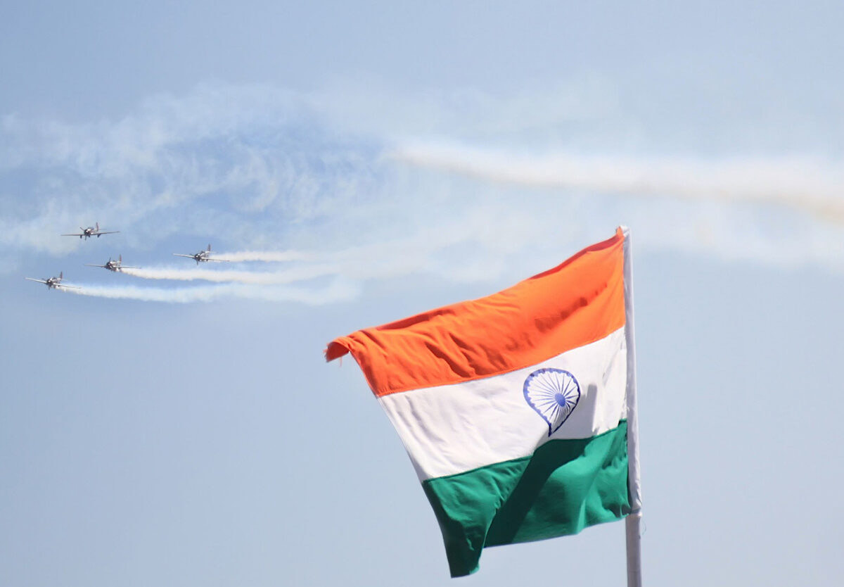 A national flag flying against the sky with a formation of aircraft trailing smoke behind it