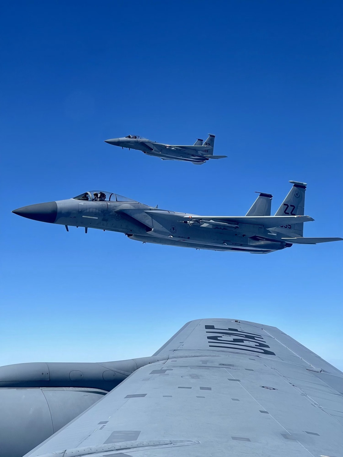 Two fighter aircraft flying in formation alongside the wing of a larger air force aircraft