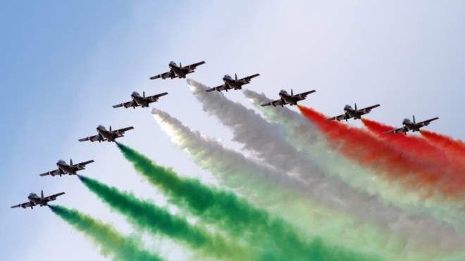 A formation of aircraft flying in close order trailing green, white and orange smoke